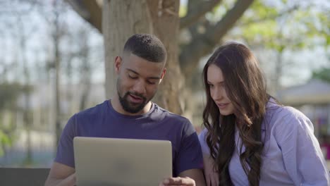 Young-man-and-pretty-woman-in-park-looking-at-laptop,-discussing